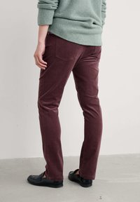 Burgundijas krāsas corduroy bikses ar šauru piegriezumu, ar aizmugurējām kabatu detaļām, apvienotas ar melniem loafers, uz neitrāla fona.