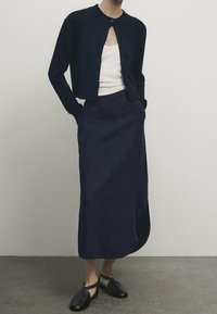 Tummansininen neulecardigan, jossa on nappi etupuolella, yhdistettynä pitkähihaiseen tummansiniseen hameeseen. Hameessa on kevyt sivuhaalari ja se on tiukka muotoilultaan. Mustat liukukengät.