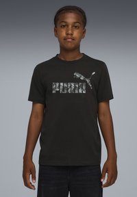 Musta puuvillapaita, jossa on harmaa camo "PUMA" -logo ja hyppivä pumagrafiikka. Pyöreä kaula-aukko, lyhyet hihat ja rento leikkaus.