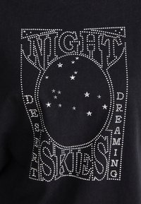Juodas marškinėlis su sidabrinėmis studijomis užrašais "NIGHT SKIES" ir "DREAMING", su centriniu ovaliu dizainu ir žvaigždžių puošmenomis.