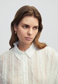 Chemise blanche avec des franges texturées, fermeture à boutons et col classique. Le tissu est transparent et superposé avec des rayures horizontales.