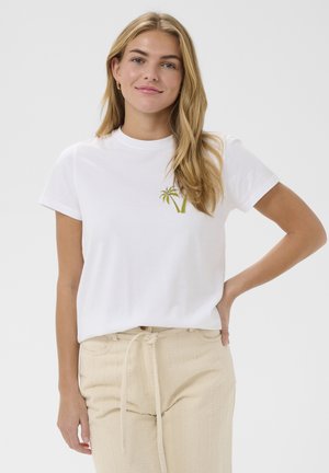 Junge Frau mit langem blondem Haar trägt ein weißes T-Shirt mit grüner Palme-Stickerei und beige Hosen, steht mit einer Hand an der Hüfte.