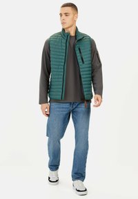 Grüne gesteppte Weste mit hohem Kragen, Reißverschluss vorne und schwarzen Akzenten, getragen über einem grauen Langarmshirt und blauen Jeans. Legere Sneakers.