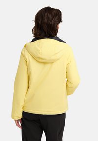 Giacca gialla impermeabile con cappuccio e dettagli neri, caratterizzata da una texture liscia e una vestibilità larga, con cuciture visibili lungo le giunture.