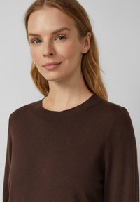 Blondynka z niebieskimi oczami, ubrana w ciemno-brązowy sweter z okrągłym dekoltem, stojąca na tle jasnoszarej ściany, delikatnie uśmiechająca się.