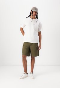 Hvid bomulds t-shirt med trykt tekst, olivengrøn cargo shorts, grå kasket og hvide sneakers med sorte accenter.