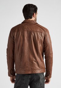 Bruine leren jas met doorgestikte schouderdetails, getailleerd ontwerp en drukknoopsluiting. Draagt over donkere spijkerbroek. Gladde textuur.