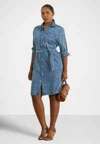 Žena v zapínacej rifľovej košeľovej sukni s vyhrnutými rukávmi a viazaným pásikom v páse, drží hnedú kabelku a má na nohách hnedé sandále s remienkom okolo členka.