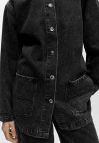 Zwarte denim jas met een knoopsluiting aan de voorkant, twee zijzakken en subtiele stiksel details. De stof heeft een gestructureerde afwerking.