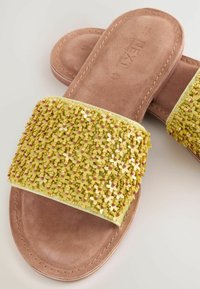 Sandalias planas con una tira de cuentas amarillas que presenta acentos en forma de cruz, sobre una plantilla de gamuza marrón. Textura suave y diseño abierto.