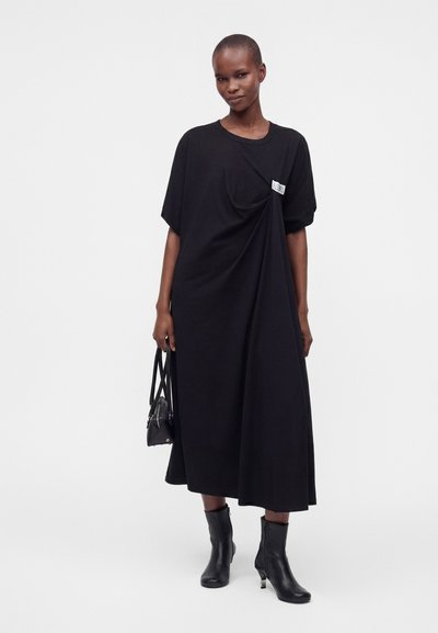 Grande femme portant une robe midi noire ample avec une petite étiquette blanche, des bottines noires, tenant un sac à main noir sur fond blanc.