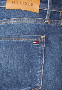 Jeans de denim en un color azul oscuro con costuras en contraste, etiqueta de cuero y un pequeño logo en rojo, blanco y azul en el bolsillo trasero.