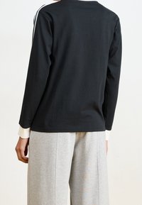 Osoba oblečená v černém tričku s dlouhým rukávem s bílými pruhy na ramenou a šedých kalhotách s širokými nohavicemi, zobrazená zezadu.