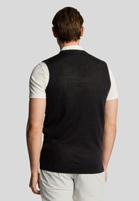 Gilet in maglia nero senza maniche realizzato in materiale leggero, design aderente, con scollatura e orlo a costine, indossato sopra una camicia bianca.