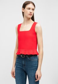 Mujer con una blusa sin mangas roja acanalada con dobladillo con volantes y jeans azules, de pie contra un fondo claro y liso.