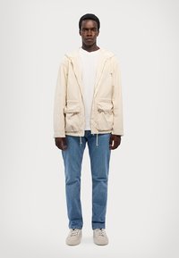 Svetlo bež jakna s kapuco, z dvema sprednjima žepoma, nanešena čez belo majico. Ujemanje z svetlo modrimi kavbojkami in bež supergami.