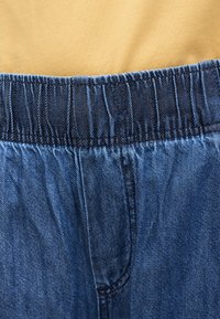 Κοντινό πλάνο της ελαστικής μέσης σε μπλε τζιν παντελόνια που φοριούνται με μπλούζα σε μουσταρδί απόχρωση, παρουσιάζοντας την υφή του υφάσματος και τις ραφές.