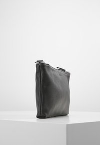 Pochette en cuir noir avec fermeture éclair, placée verticalement sur un socle blanc, devant un fond blanc uni.