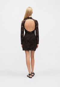 Robe noire à manches longues avec un voile floral transparent, découpe dans le dos et taille cintrée. Elle présente une fermeture à nouer au niveau du cou et une jupe évasée.