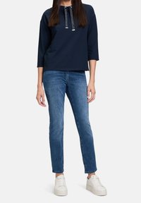 Sudadera marinera con escote redondo, mangas largas y un cordón plateado. Combinada con unos vaqueros ajustados de color azul claro y zapatillas blancas.