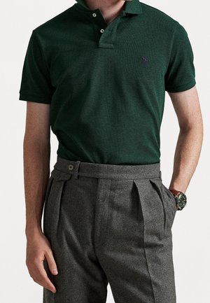 Homme portant un polo vert foncé à manches courtes rentré dans un pantalon gris plissé taille haute, avec une montre au poignet gauche.