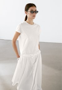 Hvid bomulds t-shirt parret med en lang, luftig hvid nederdel. Outfittet har en elastisk talje og en afslappet pasform. Sorte, ovale solbriller fuldender looket.