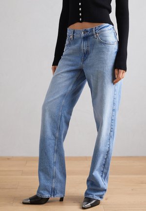 Vrouw met lichtblauwe jeans met rechte pijpen, zwarte laarzen met puntige neuzen en metalen tips, en een zwart kortgebreid vest met knopen.