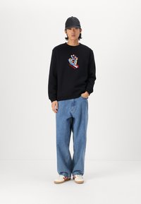 Felpa nera con grafica colorata sul petto, abbinata a jeans a gamba larga blu chiaro e sneaker bianche con accenti rossi.