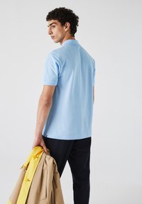 Polo shirt bleu clair à manches courtes et col, associé à un pantalon sombre. Le modèle tient une veste de pluie jaune dans une main.