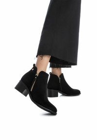 Botas de tobillo de ante negro con tacón cuadrado, que cuentan con cremalleras laterales y punta redondeada, complementadas por una textura suave y mate.