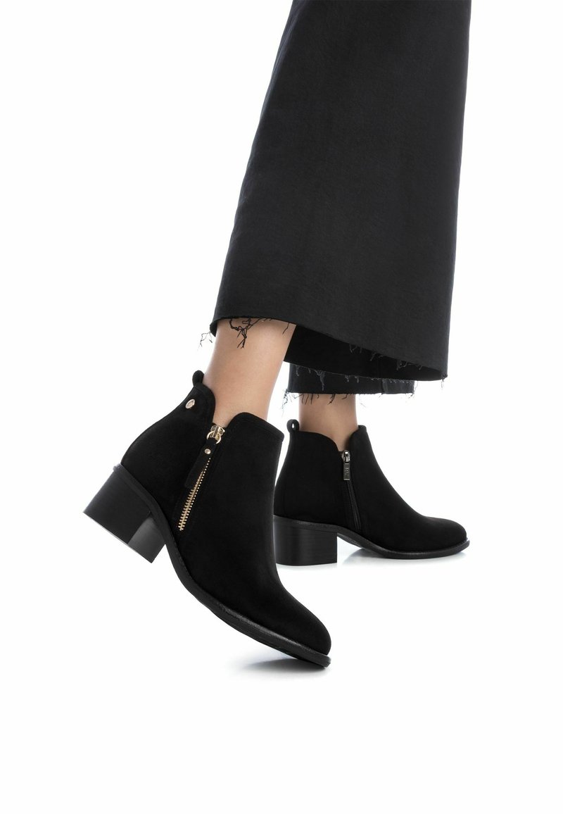 Botas de tobillo de ante negro con tacón cuadrado, que cuentan con cremalleras laterales y punta redondeada, complementadas por una textura suave y mate.