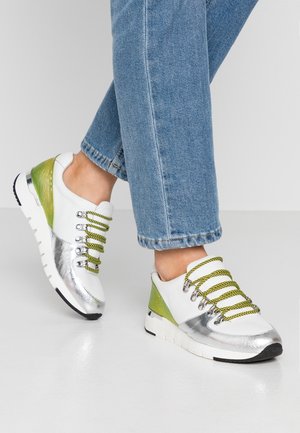 Person iført blå jeans og hvide sneakers med sølv- og grønne accenter, med gule og sorte stribede snørebånd, stående på et hvidt gulv.