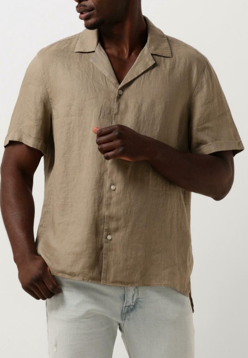 Uomo che indossa una camicia a maniche corte marrone chiaro con bottoni e jeans azzurri, con una mano vicino al petto e l'altra in tasca.