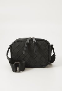 Compact zwarte crossbody tas met geweven textuur, dubbele ritshandles en verstelbare schouderband met metalen gesp op een effen achtergrond.