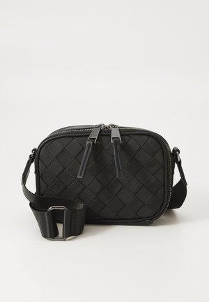 Kompakti musta crossbody-laukku, jossa on kudottu pinta, kaksoisvetoketjun vetimät ja säädettävä hihna metallisoljella yksinkertaisella taustalla.