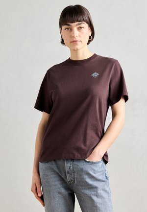 Brown cotton t-shirt with a relaxed fit, featuring a small blue graphic design on the chest. Paired with light blue denim jeans.