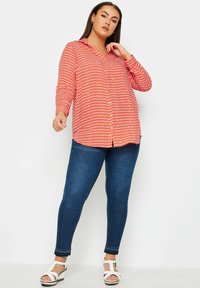 Camicia a bottoni rossa a fantasia con maniche lunghe, abbinata a jeans skinny blu scuro e sandali con zeppa bianchi.