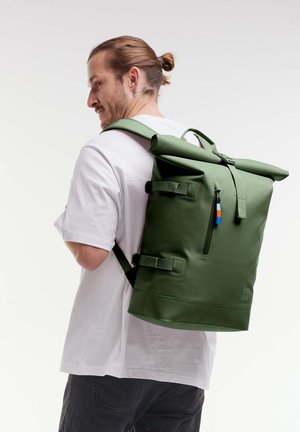 Zaino verde a sacca con cinturini regolabili, dotato di una tasca frontale con zip e fibbie laterali. Materiale liscio e impermeabile con finitura opaca.