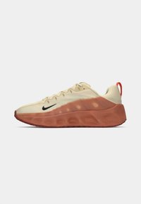 Light beige sneaker with a textured red outsole, black logo detail, and a red pull tab at the heel. Features a cushioned, breathable design.