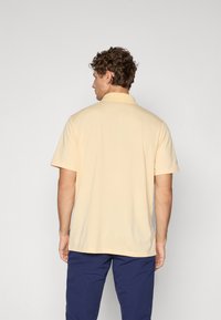 Gul polo skjorte med krage, korte ermer og rett nederkant, laget av glatt stoff, iført med marineblå bukser.