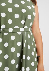 Grünes Kleid mit großen weißen Polka-Dots, mit tailliertem Schnitt und schwingendem Rock. Die strukturierte Stoffqualität sorgt für Tiefe und visuelles Interesse.