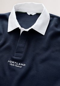 Tumesinine polo-särk valge krae, soonilise tekstuuriga ja tikitud "PORTLAND Pacific Northwest" detailiga. Nööbitav plastron.