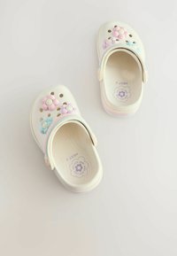 Zoccoli per bambini color crema con decorazioni di fiori pastello e cinturini a righe arcobaleno su una superficie chiara e uniforme.