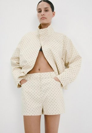 Mujer con chaqueta de manga larga color crema y pantalones cortos a juego con pequeños lunares negros, con las manos en los bolsillos, de pie frente a un fondo blanco.