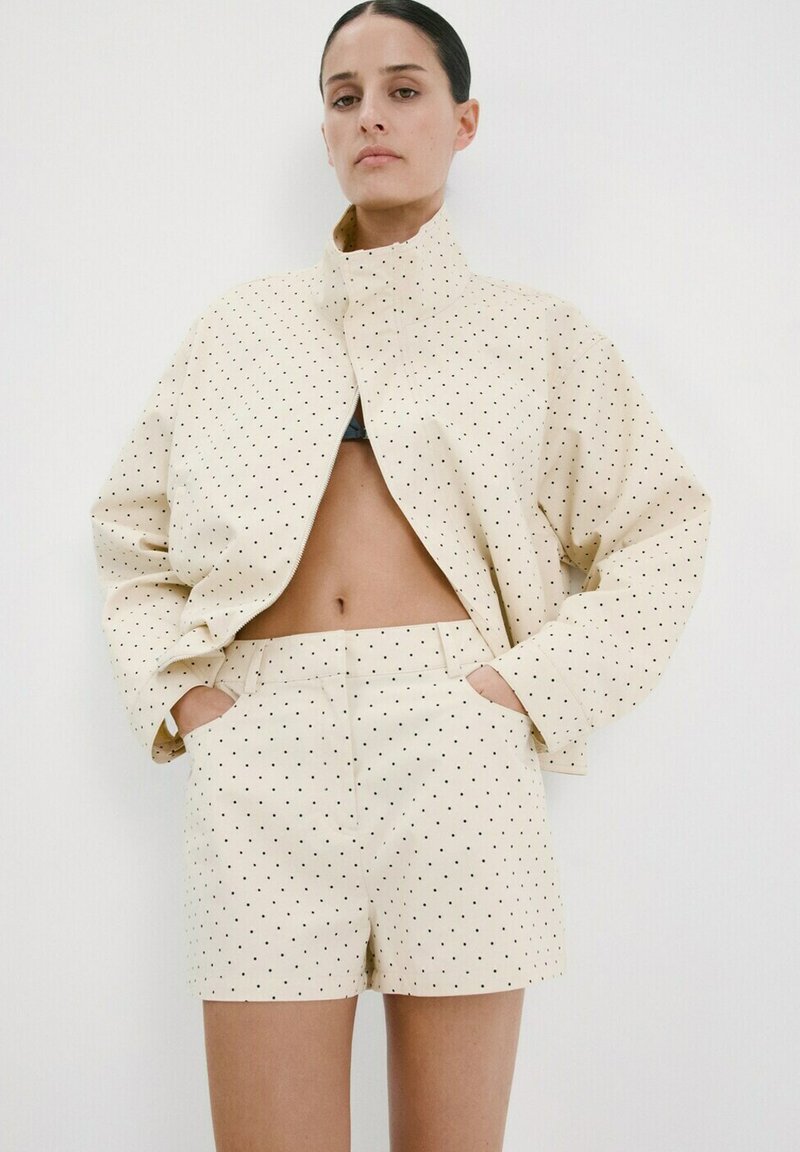 Femme portant une veste crème à manches longues et un short assorti avec de petits pois noirs, les mains dans les poches, debout devant un fond blanc.