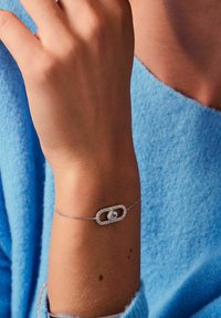 Bracciale d'argento con un pendente rettangolare caratterizzato da una pietra centrale, circondata da piccoli accenti decorativi. Indossato su un polso.