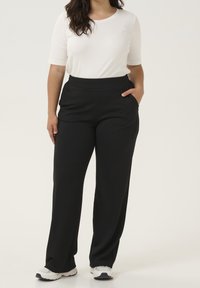 Pantalons noirs à jambes larges avec une texture lisse et une taille élastique, associés à un haut ajusté à col rond blanc. L'utilisateur a les mains dans les poches.