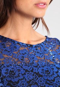 Sininen pitsitoppi, jossa on kukkakuvioita ja upotettuja paljetteja, erottuva musta vuoraus ja pyöristetty pääntie, joka luo teksturoidun lopputuloksen.