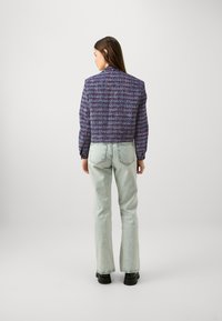 Geknipte, gestructureerde jas in paars met meerkleurige patronen, gecombineerd met lichte denim flarebroeken en zwarte schoenen.