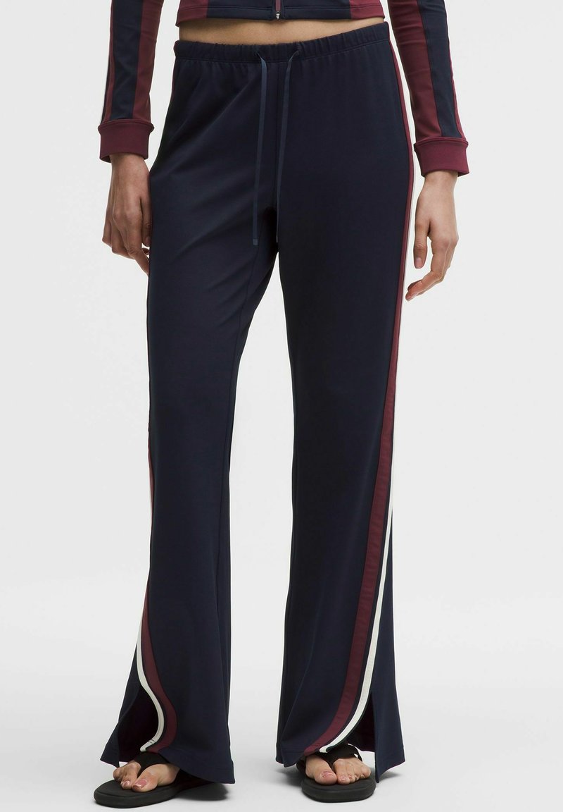 Person trägt marineblaue, weit geschnittene Hosen mit bordeauxroten und weißen Seitenstreifen, elastischem Bund mit Kordelzug und schwarzen Sandalen.